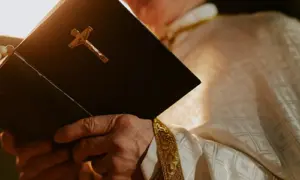 Symbolbild: Ein Geistlicher im liturgischen Gewand hält eine geschlossene Bibel mit goldenem Kruzifix in den Händen