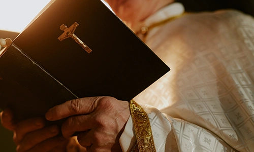 Symbolbild: Ein Geistlicher im liturgischen Gewand hält eine geschlossene Bibel mit goldenem Kruzifix in den Händen
